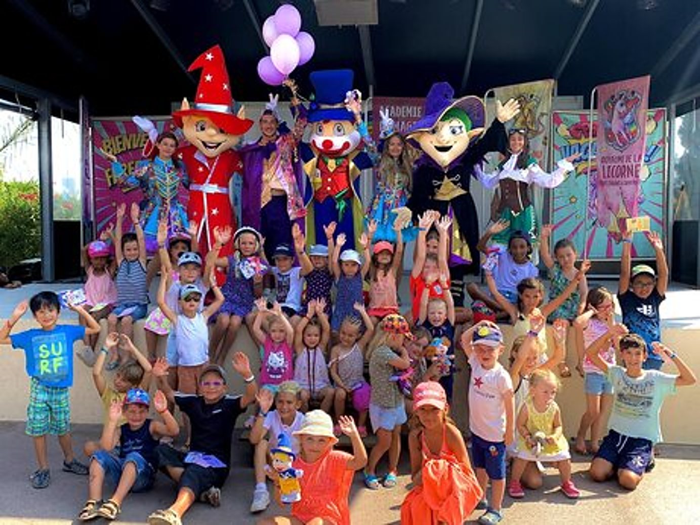 Un grand groupe d'enfants lève les bras en posant devant des mascottes costumées et des animatrices sur une scène en plein air.