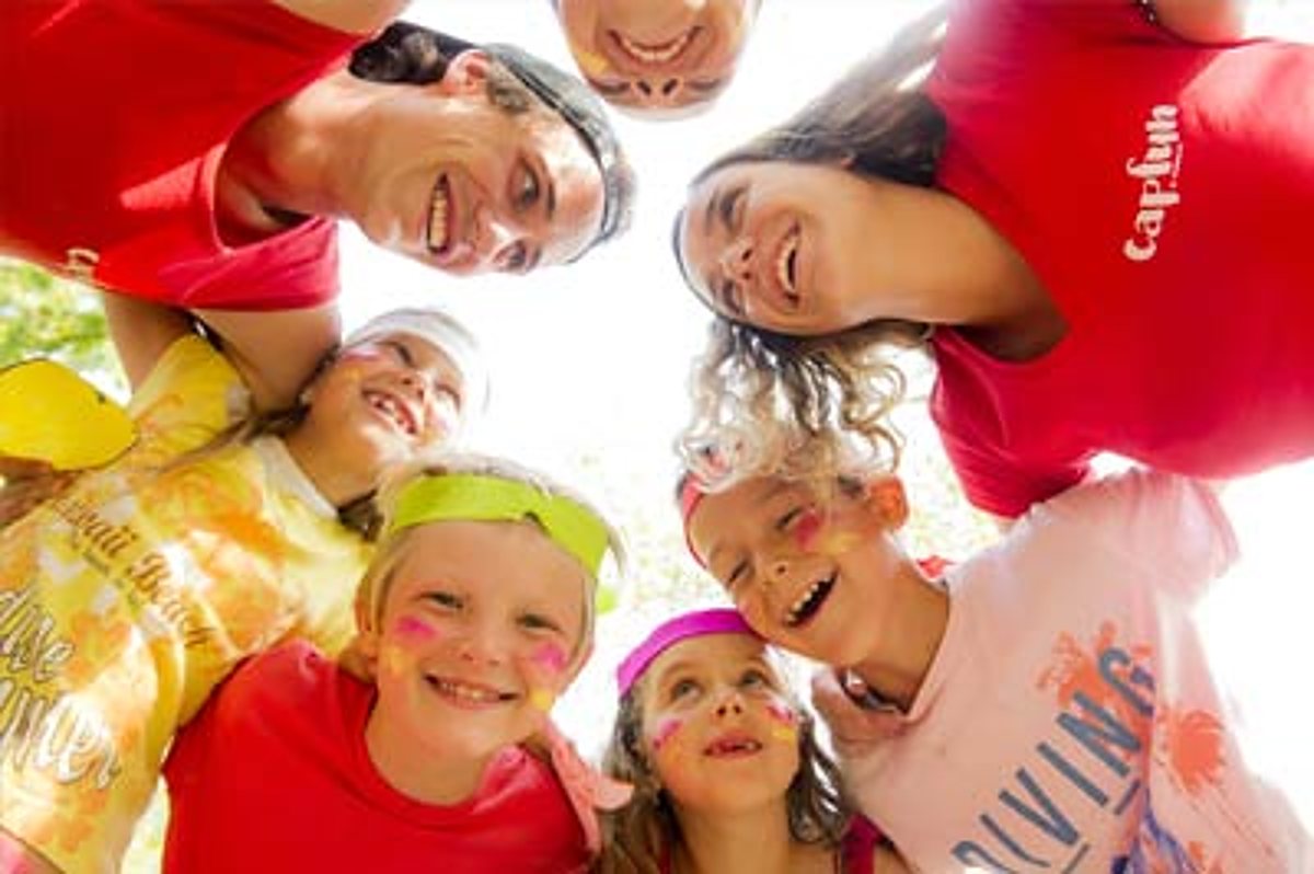 Des enfants et des adultes en t-shirts colorés forment un cercle et sourient à la caméra, photographiés depuis le bas.