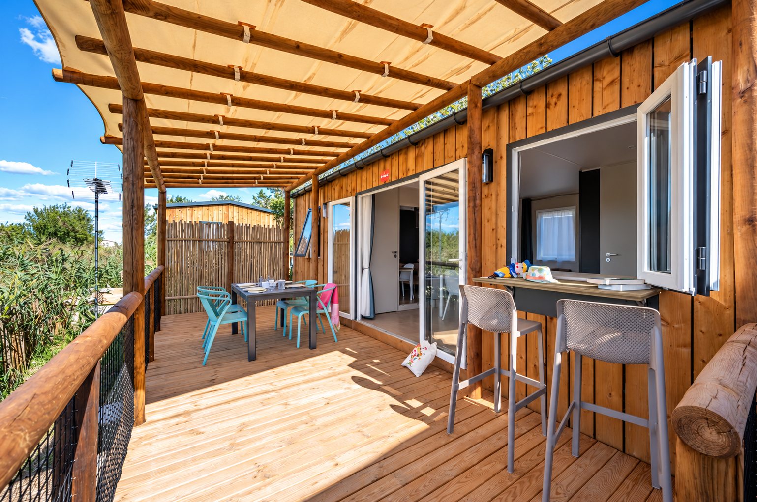 Mobil-home en bois avec terrasse au camping Fleur de Camargue : terrasse sous pergola avec table, chaises turquoise et tabourets hauts.