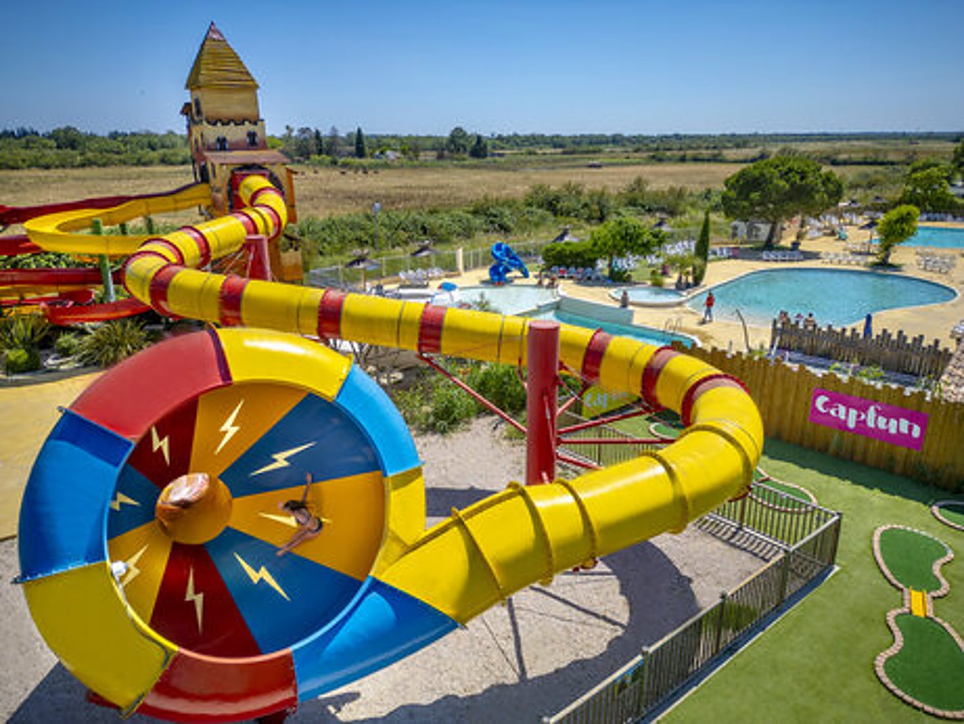 Vue aérienne de grands toboggans jaunes et colorés avec des piscines et un mini-golf dans un parc aquatique sous un ciel bleu.