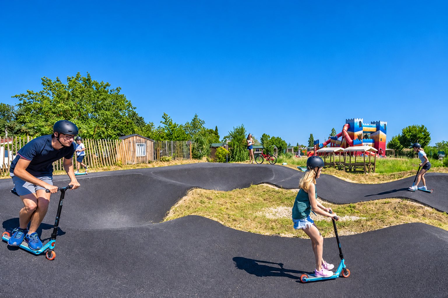 Un homme et une jeune fille en trottinette roulent sur une piste de pumptrack en enrobé noir, sous un ciel bleu, avec une structure gonflable en arrière-plan.